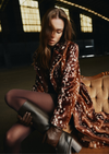 A woman sits on a tufted sofa in a dimly lit interior, wearing a brown faux-fur coat with a white spotted pattern, dark tights, and tall dark boots.