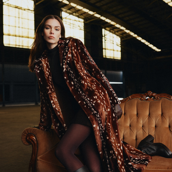 A woman sits on an ornate tan leather sofa in a dimly lit industrial loft, wearing a long brown faux-fur coat with white spotted patterning, a black dress, sheer black tights, and black knee-high boots.