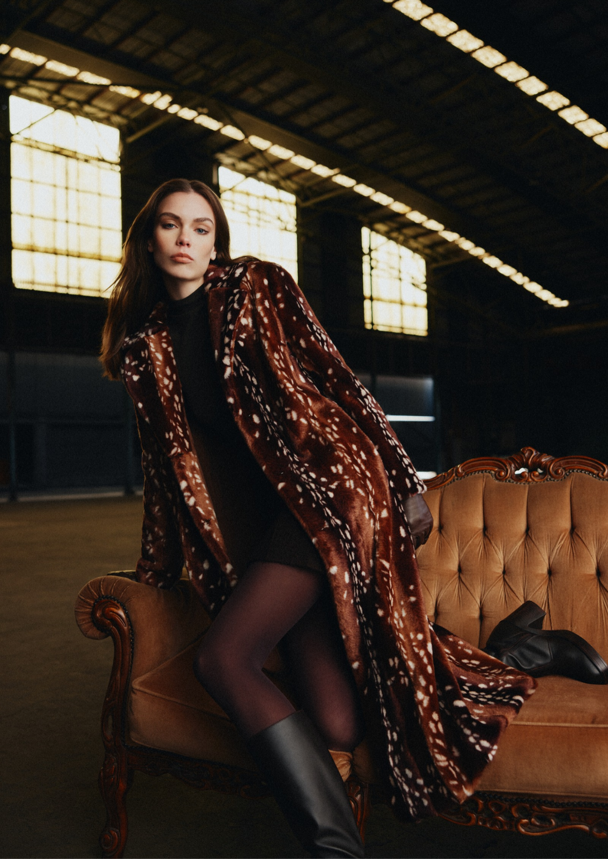 A woman sits on an ornate tan leather sofa in a dimly lit industrial loft, wearing a long brown faux-fur coat with white spotted patterning, a black dress, sheer black tights, and black knee-high boots.