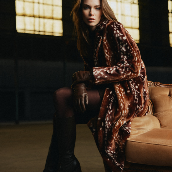 A woman sits on a tufted sofa in a dimly lit interior, wearing a brown faux-fur coat with a white spotted pattern, dark tights, and tall dark boots.