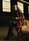 A woman sits on a tufted sofa in a dimly lit interior, wearing a brown faux-fur coat with a white spotted pattern, dark tights, and tall dark boots.