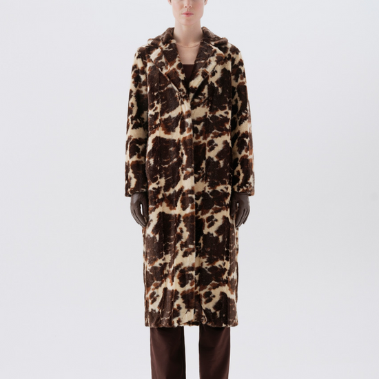 A model stands facing forward wearing a long brown and beige animal-print faux fur coat, dark brown trousers, and black boots against a light gray studio background.