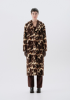 A model stands facing forward wearing a long brown and beige animal-print faux fur coat, dark brown trousers, and black boots against a light gray studio background.