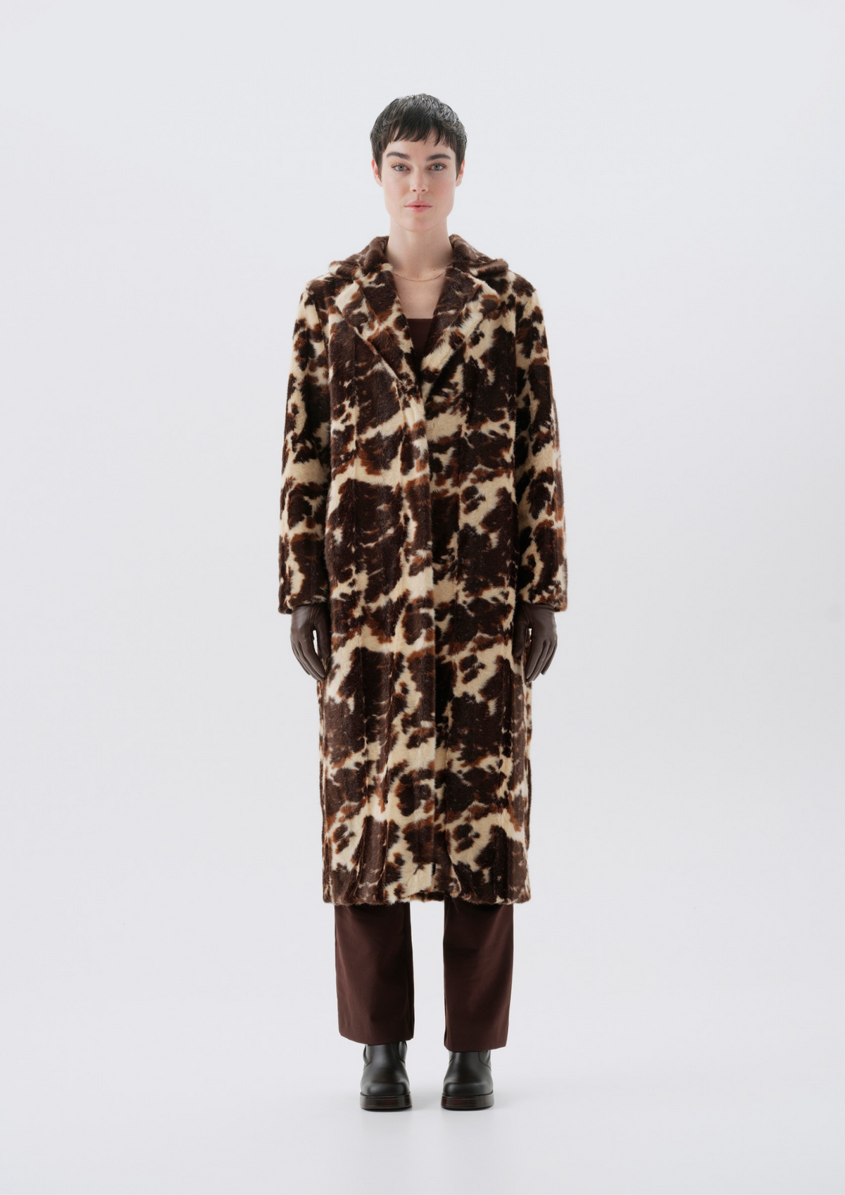 A model stands facing forward wearing a long brown and beige animal-print faux fur coat, dark brown trousers, and black boots against a light gray studio background.
