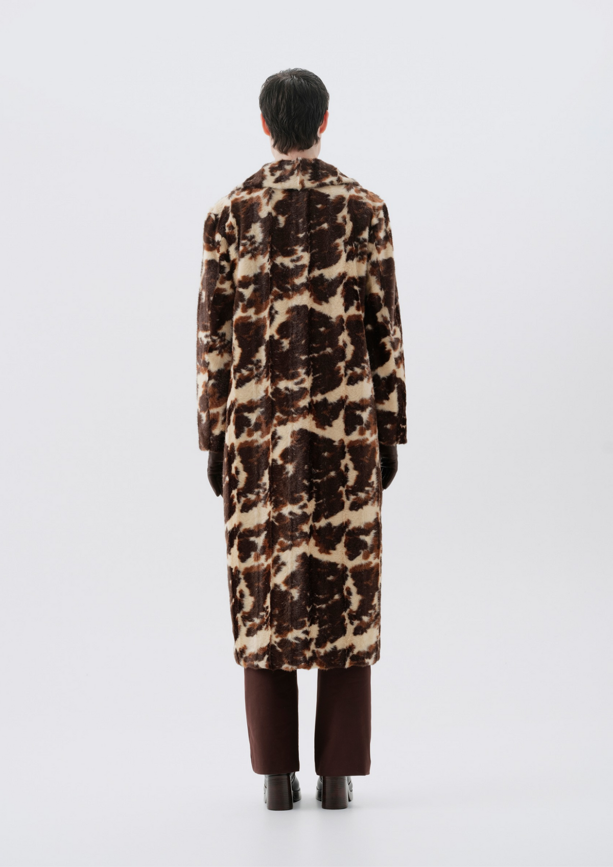 A model viewed from the back wearing a long brown and beige animal-print faux fur coat, paired with dark brown trousers and black shoes, against a light gray studio background.