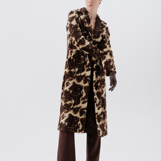 A model wearing a long brown and beige animal-print coat, dark brown trousers, and black boots, standing against a plain light gray studio background.