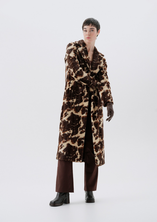 A model wearing a long brown and beige animal-print coat, dark brown trousers, and black boots, standing against a plain light gray studio background.