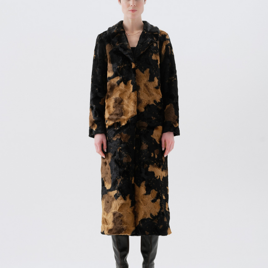 A model stands facing forward in a studio wearing a long black and brown abstract patterned coat over a black outfit with black boots, against a light gray background.