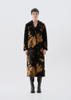 A model stands facing forward in a studio wearing a long black and brown abstract patterned coat over a black outfit with black boots, against a light gray background.