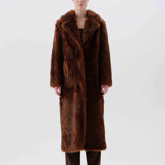 A model stands facing forward wearing a long, plush brown fur coat with a wide collar, paired with dark brown pants and black boots, against a seamless light gray studio background.