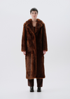 A model stands facing forward wearing a long, plush brown fur coat with a wide collar, paired with dark brown pants and black boots, against a seamless light gray studio background.