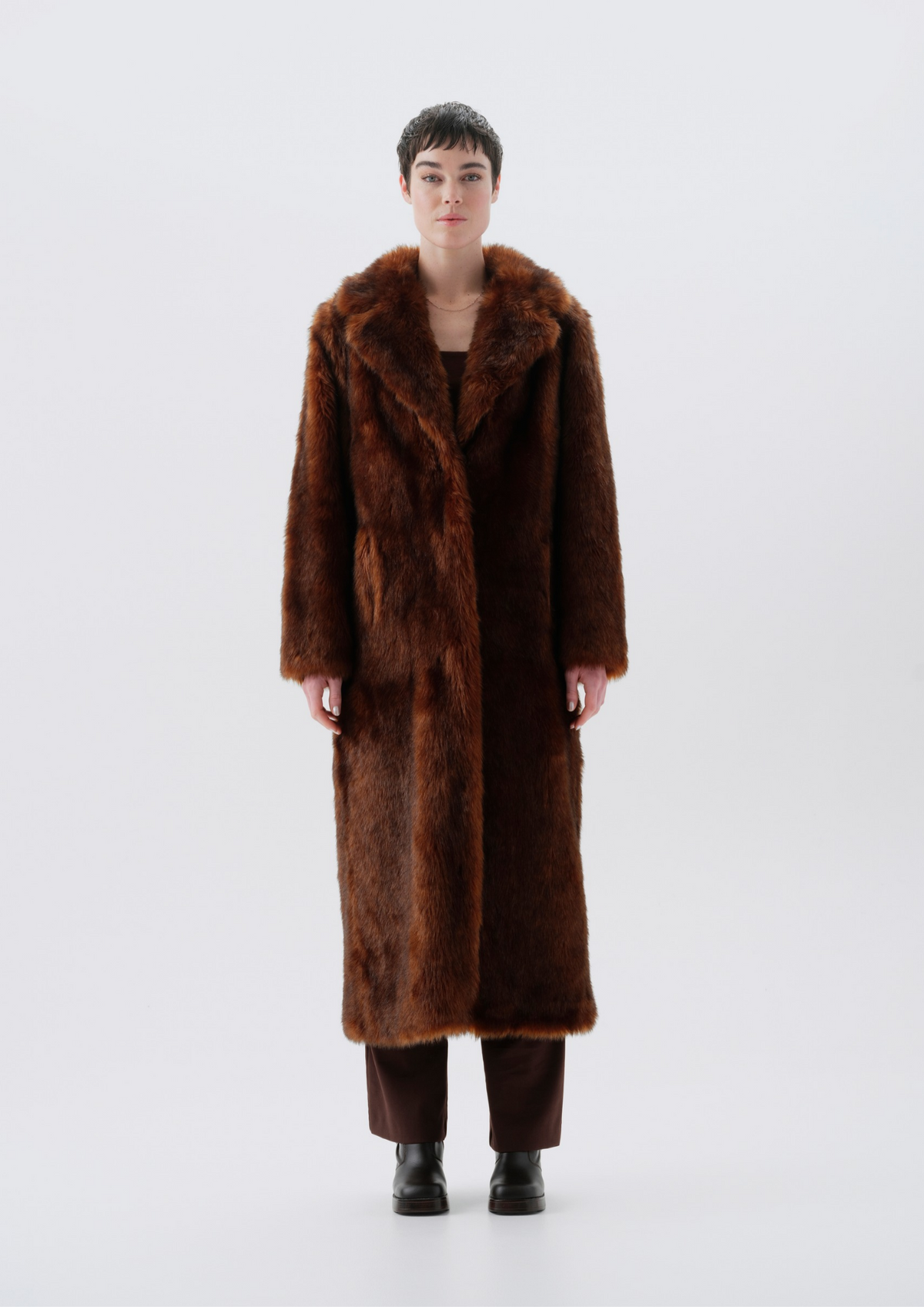 A model stands facing forward wearing a long, plush brown fur coat with a wide collar, paired with dark brown pants and black boots, against a seamless light gray studio background.