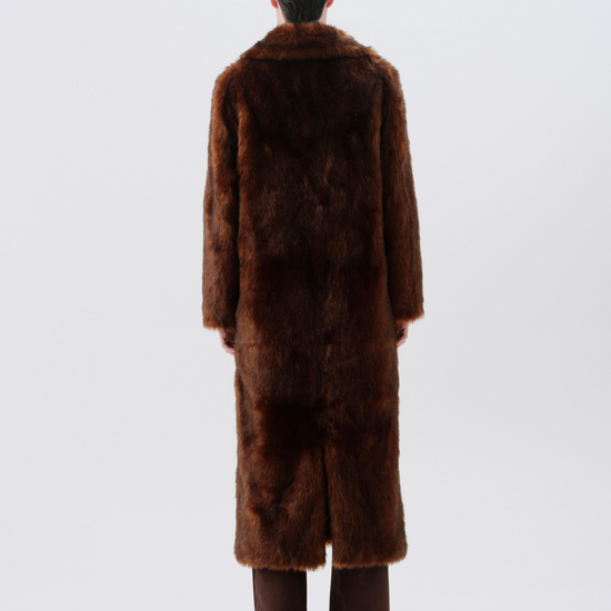A model viewed from behind wearing a long, dark brown fur coat, brown trousers, and black shoes against a plain light gray studio background.