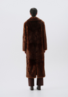 A model viewed from behind wearing a long, dark brown fur coat, brown trousers, and black shoes against a plain light gray studio background.