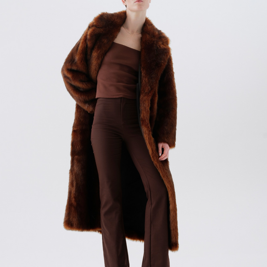 A model stands against a light gray studio background wearing a long brown faux-fur coat over a matching brown strapless top and brown trousers, with black ankle boots.