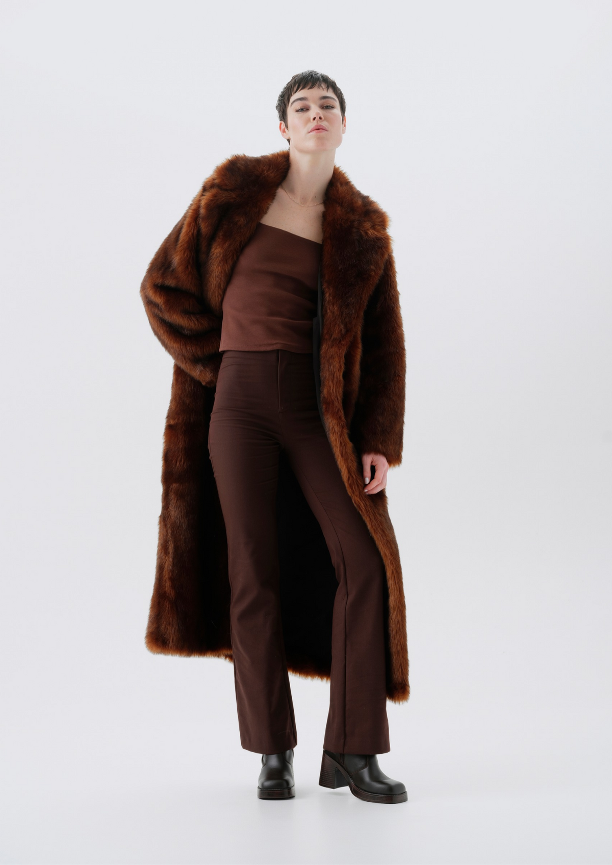 A model stands against a light gray studio background wearing a long brown faux-fur coat over a matching brown strapless top and brown trousers, with black ankle boots.