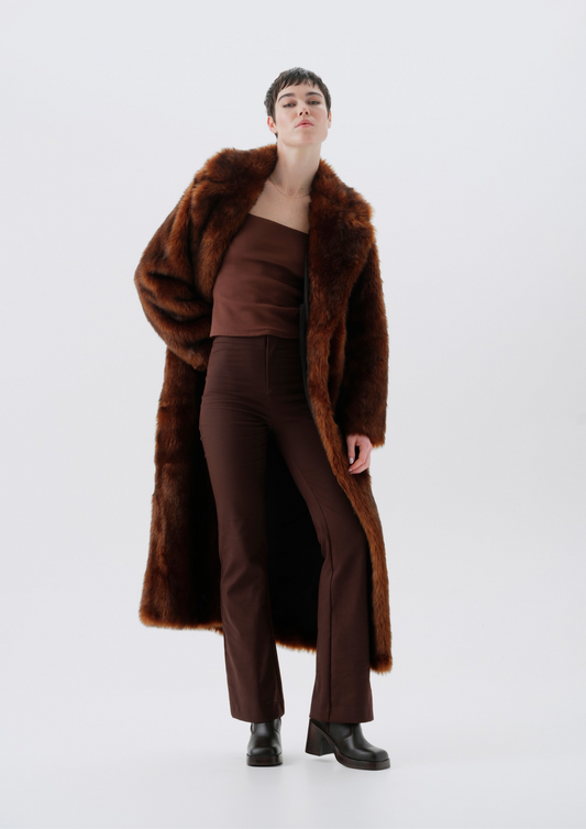 A model stands against a light gray studio background wearing a long brown faux-fur coat over a matching brown strapless top and brown trousers, with black ankle boots.