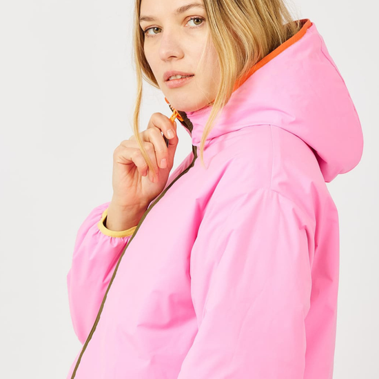 A woman wearing a bright pink hooded jacket with an orange-lined hood and a gold zipper, shown in a studio setting against a light gray background.