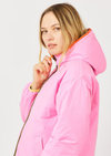 A woman wearing a bright pink hooded jacket with an orange-lined hood and a gold zipper, shown in a studio setting against a light gray background.