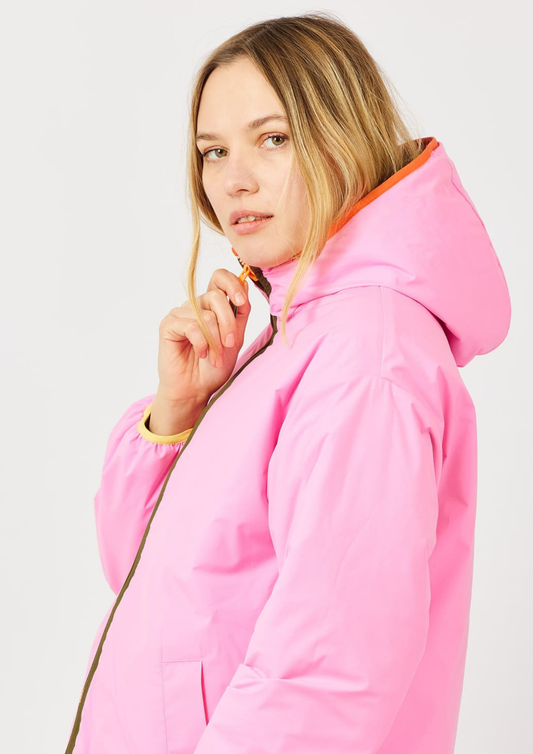 A woman wearing a bright pink hooded jacket with an orange-lined hood and a gold zipper, shown in a studio setting against a light gray background.