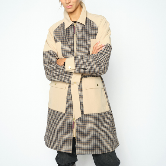 A model wearing a patchwork coat with beige and brown gingham patterns, paired with dark pants and brown leather boots, against a light gray studio background. close up