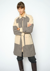 A model wearing a patchwork coat with beige and brown gingham patterns, paired with dark pants and brown leather boots, against a light gray studio background. close up