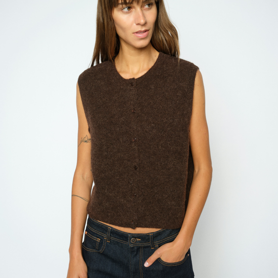 A woman modeling a sleeveless, crew-neck knit vest in a dark brown color, paired with dark blue jeans, against a plain light gray background. 