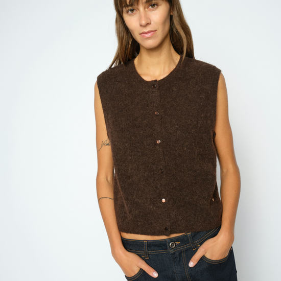 A woman modeling a sleeveless, crew-neck knit vest in a dark brown color, paired with dark blue jeans, against a plain light gray background.