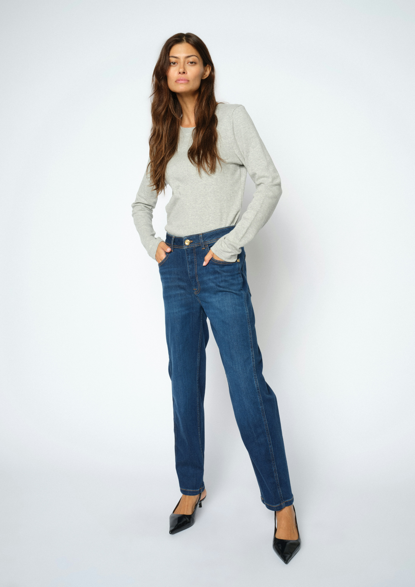 A woman modeling a light gray long-sleeve knit top and blue straight-leg jeans with black pointed-toe heels, standing against a light gray studio background.