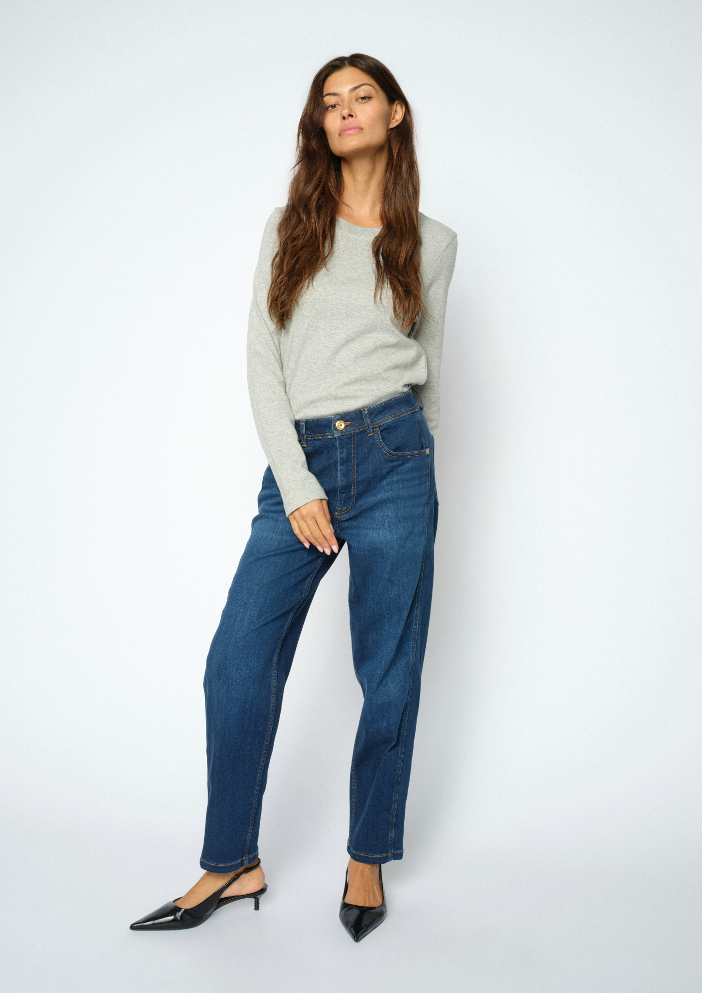 A woman modeling a light gray long-sleeve knit sweater, dark blue straight-leg jeans, and black pointed-toe slingback heels against a plain light gray studio background.