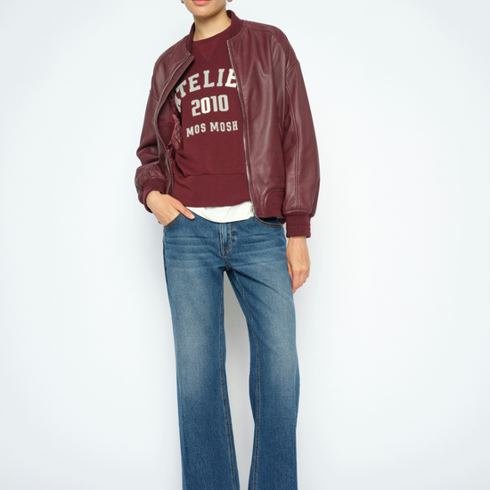 A model stands against a light gray studio background wearing a burgundy faux-leather bomber jacket over a burgundy graphic sweatshirt, blue jeans, and black boots.