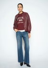 A model stands against a light gray studio background wearing a burgundy faux-leather bomber jacket over a burgundy graphic sweatshirt, blue jeans, and black boots.