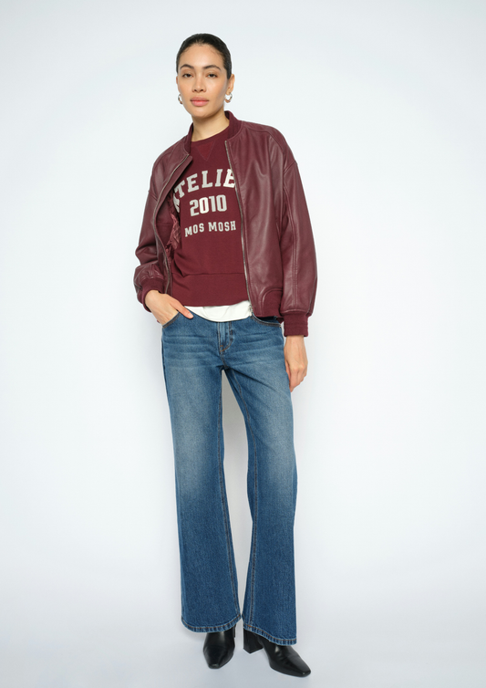 A model stands against a light gray studio background wearing a burgundy faux-leather bomber jacket over a burgundy graphic sweatshirt, blue jeans, and black boots.