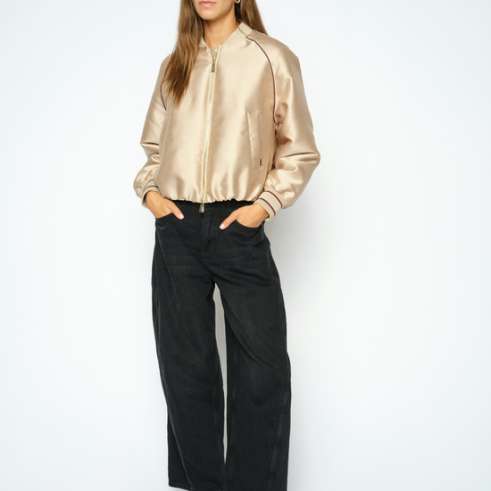 A woman modeling a shiny champagne-gold satin bomber jacket paired with black wide-leg trousers and black loafers, standing against a plain white studio background.