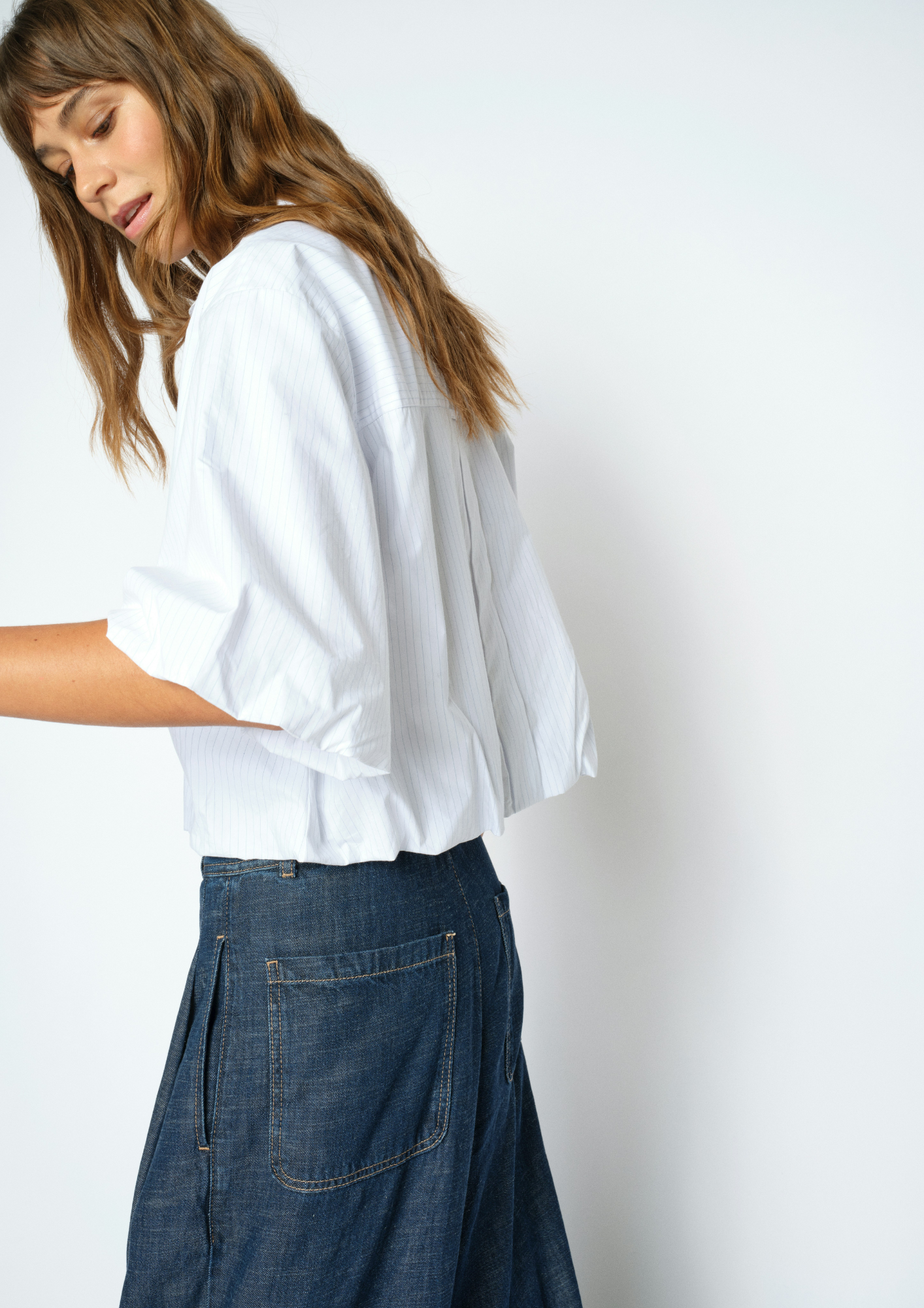 A woman wearing a white short-sleeve blouse with gathered sleeves and dark blue denim skirt, photographed against a plain white studio background.
