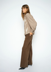 A woman stands in profile wearing a beige and brown vertically striped long-sleeve blouse with white abstract leaf-like patterns, paired with wide-leg brown trousers and black pointed-toe heels, against a plain light gray studio background.