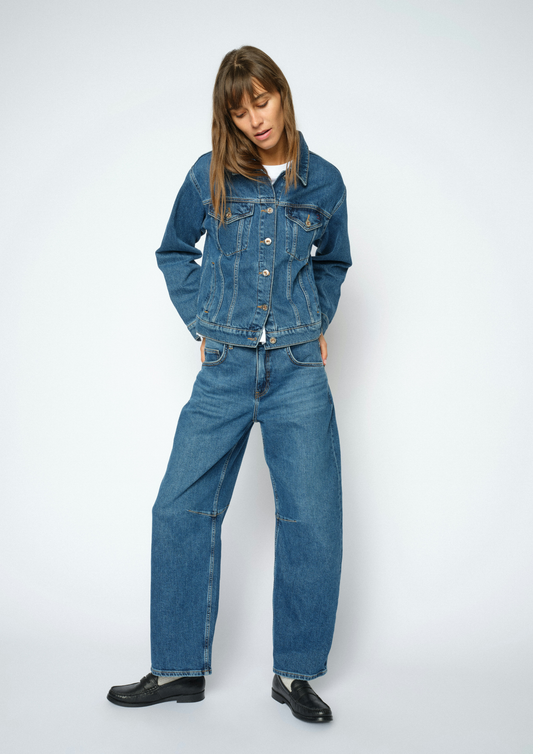 A model wearing a blue denim jacket and matching wide-leg denim jeans, styled with black leather shoes, against a light gray studio background.