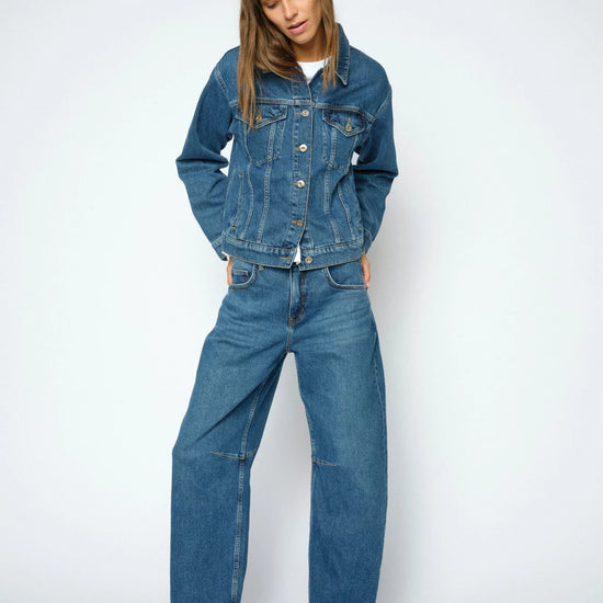 A model wearing a blue denim jacket and matching wide-leg denim jeans, styled with black leather shoes, against a light gray studio background.