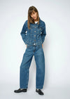 A model wearing a blue denim jacket and matching wide-leg denim jeans, styled with black leather shoes, against a light gray studio background.