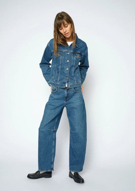 A model wearing a blue denim jacket and matching wide-leg denim jeans, styled with black leather shoes, against a light gray studio background.