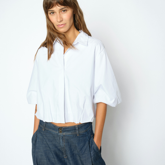 A woman modeling a white short-sleeve button-up shirt with voluminous elbow-length sleeves, paired with dark blue wide-leg jeans, against a plain light gray studio background.