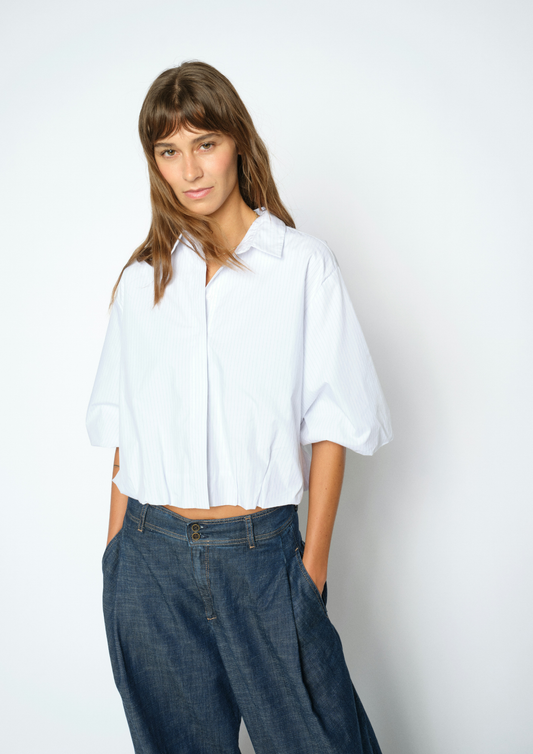 A woman modeling a white short-sleeve button-up shirt with voluminous elbow-length sleeves, paired with dark blue wide-leg jeans, against a plain light gray studio background.