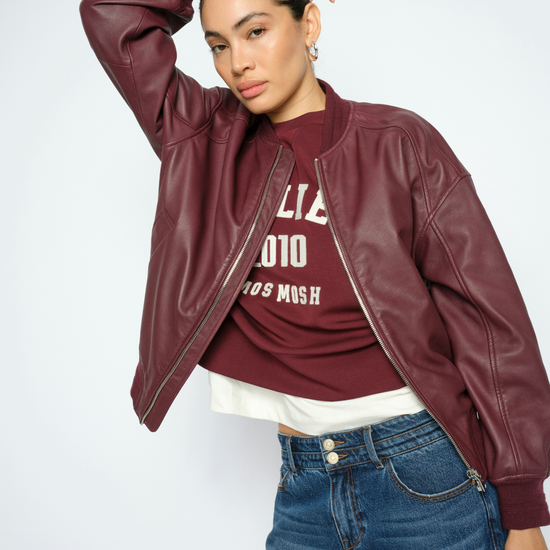A woman modeling a burgundy faux-leather bomber jacket over a matching burgundy graphic t-shirt and blue jeans, posed against a light gray studio background.