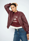 A woman modeling a burgundy faux-leather bomber jacket over a matching burgundy graphic t-shirt and blue jeans, posed against a light gray studio background.