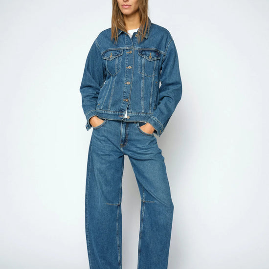 A woman modeling a matching blue denim set consisting of a cropped button-up jacket and wide-leg jeans, paired with black leather loafers, against a light gray studio background.