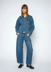 A woman modeling a matching blue denim set consisting of a cropped button-up jacket and wide-leg jeans, paired with black leather loafers, against a light gray studio background.