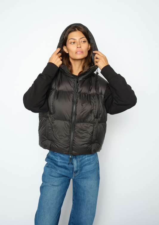 A woman wearing a black hooded puffer vest over a black long-sleeve top and blue jeans, standing against a plain light gray studio background.