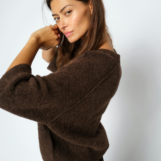 A woman wearing a fuzzy brown knit sweater and dark brown trousers, posing against a light gray studio background.