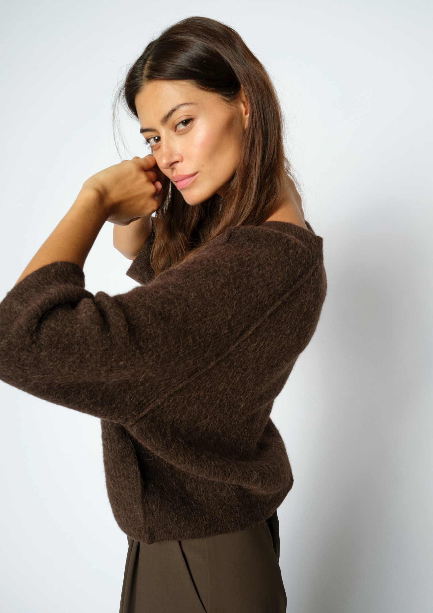 A woman wearing a fuzzy brown knit sweater and dark brown trousers, posing against a light gray studio background.