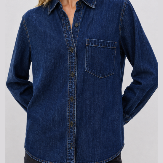 A woman wearing a dark blue denim long-sleeve button-up shirt with a chest pocket, paired with dark jeans, against a plain light gray background.